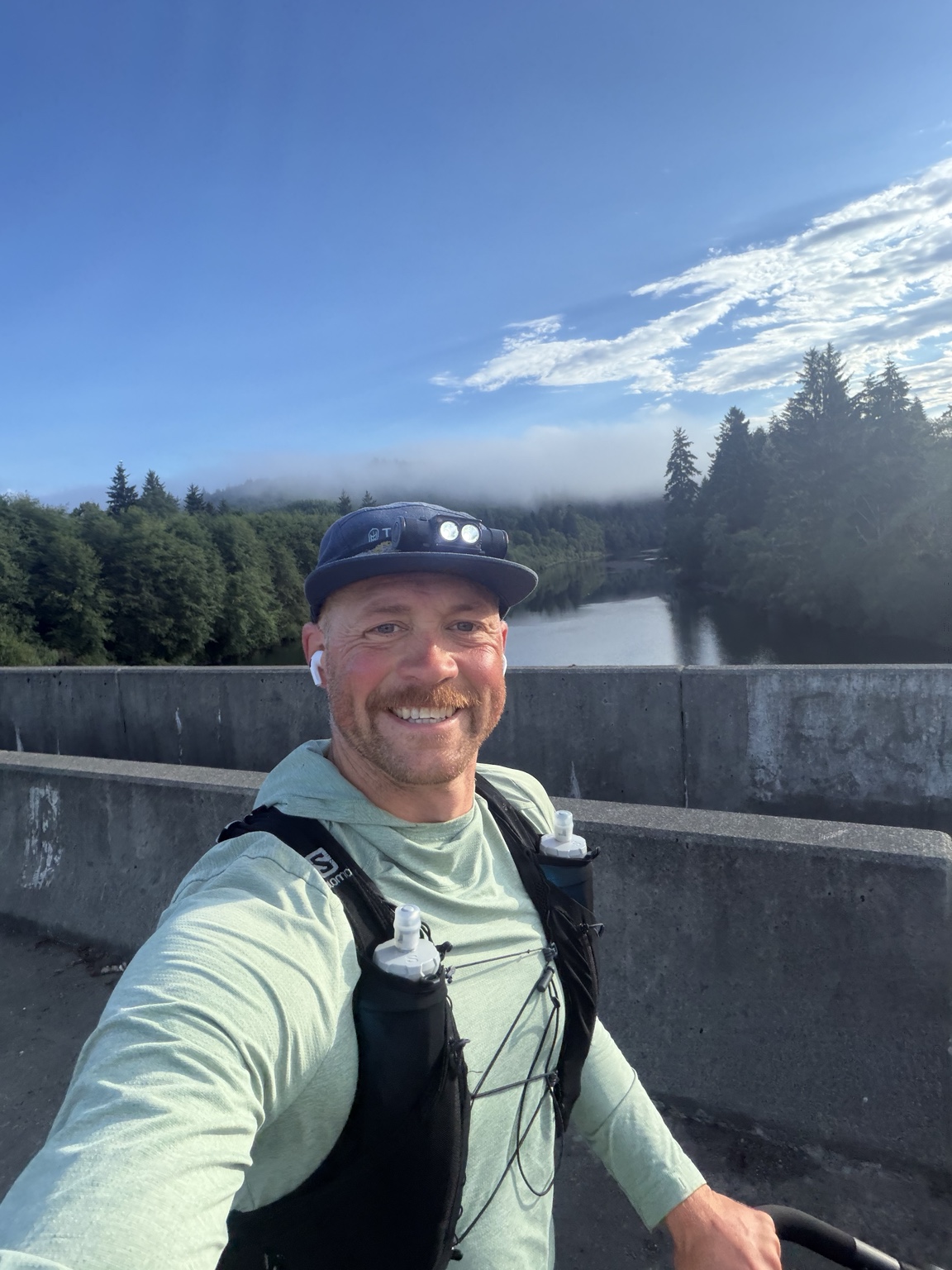 Matthew smiling on a bridge above a forest-lined river during his run to Lake Quinault