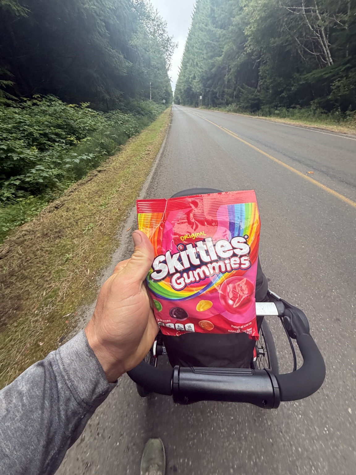Matthew holding a bag of Skittles Gummies above his stroller on a quiet forest road