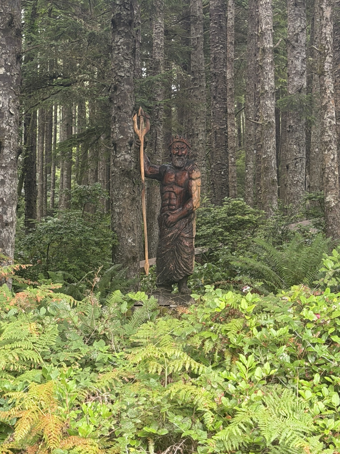 A carved figure holding a staff among ferns and trees in the coastal forest