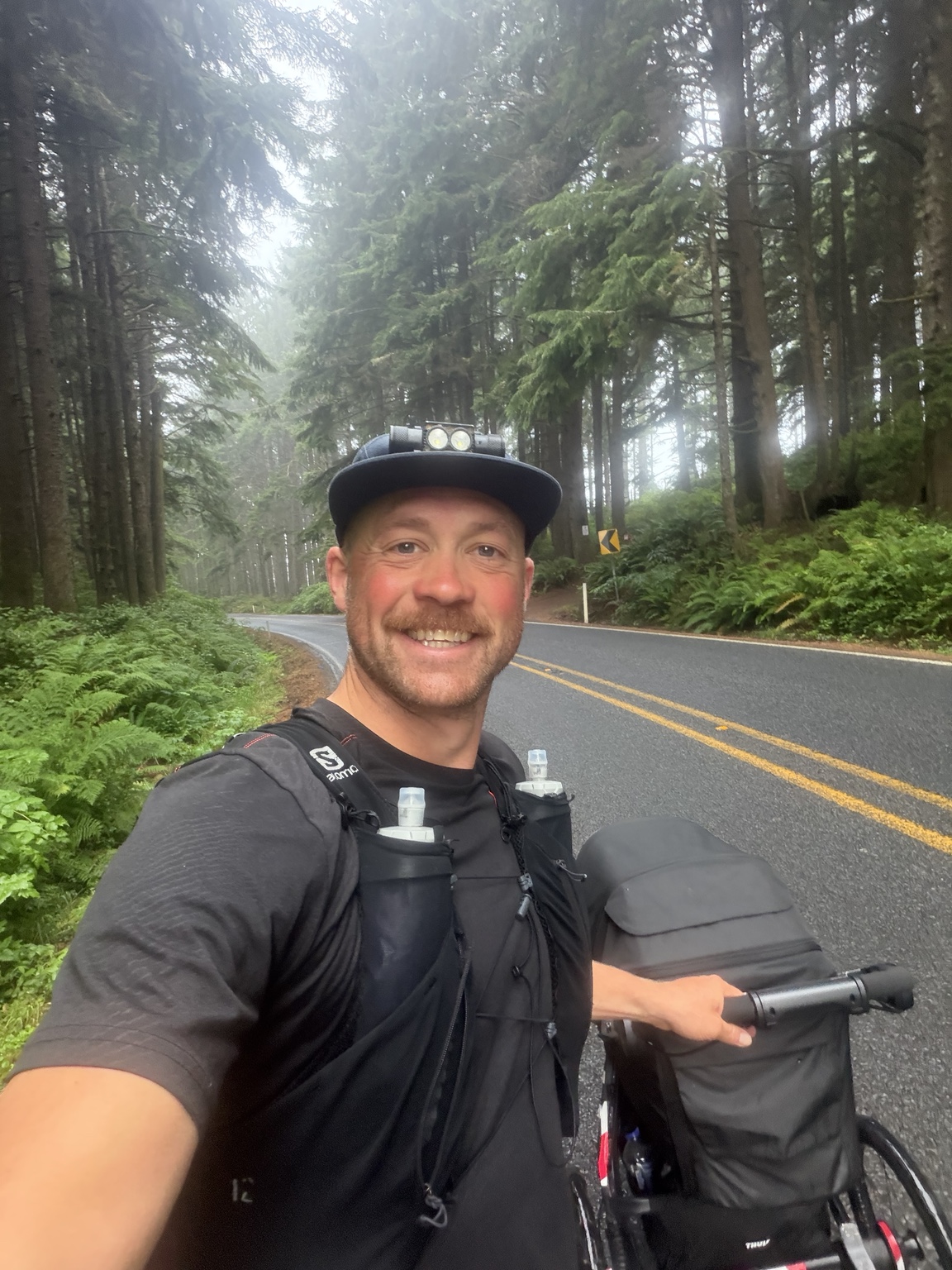 Matthew smiling beside his stroller on a misty, tree-lined coastal road