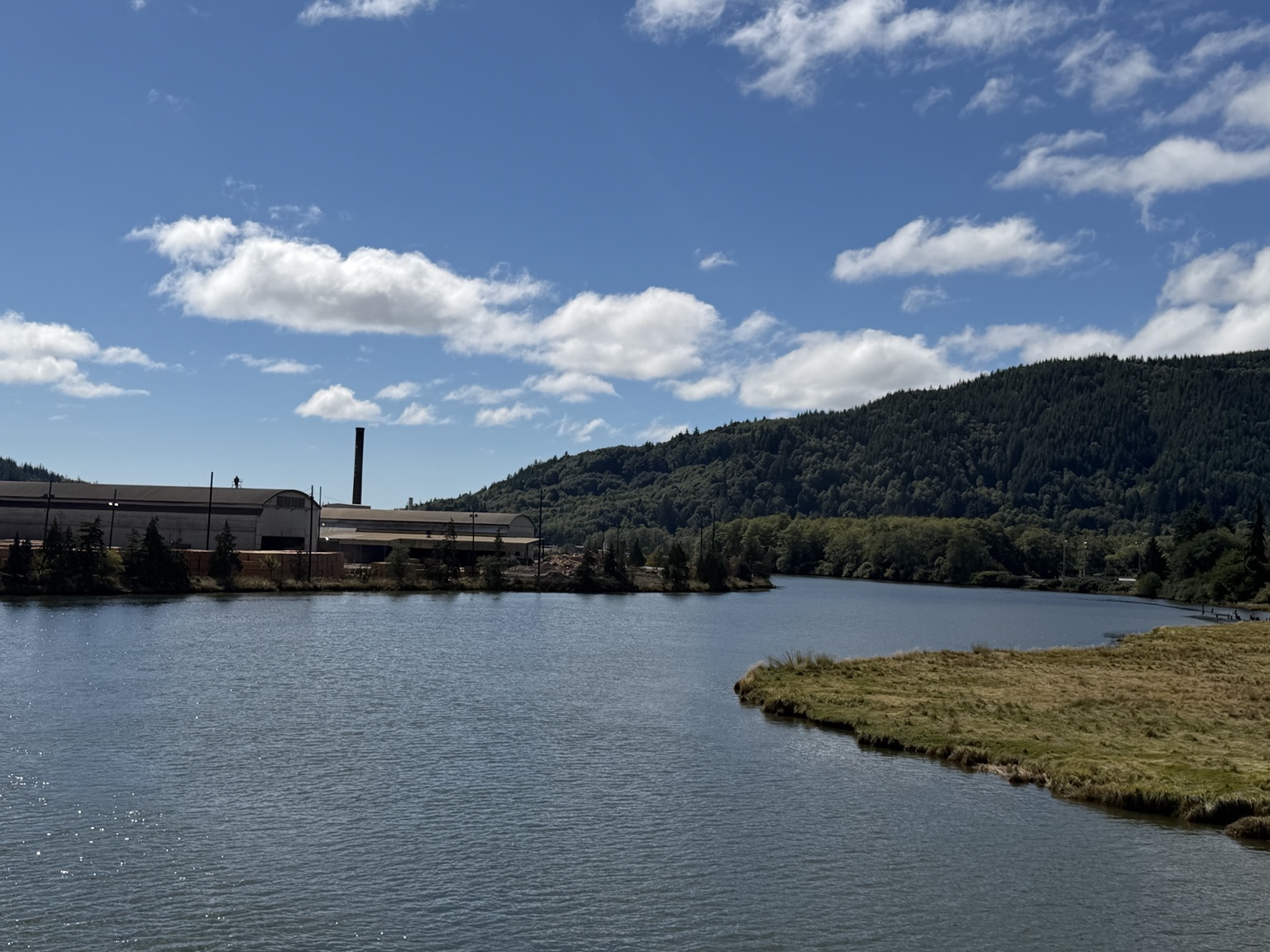 A lumber mill beside calm water and forested hills in Raymond
