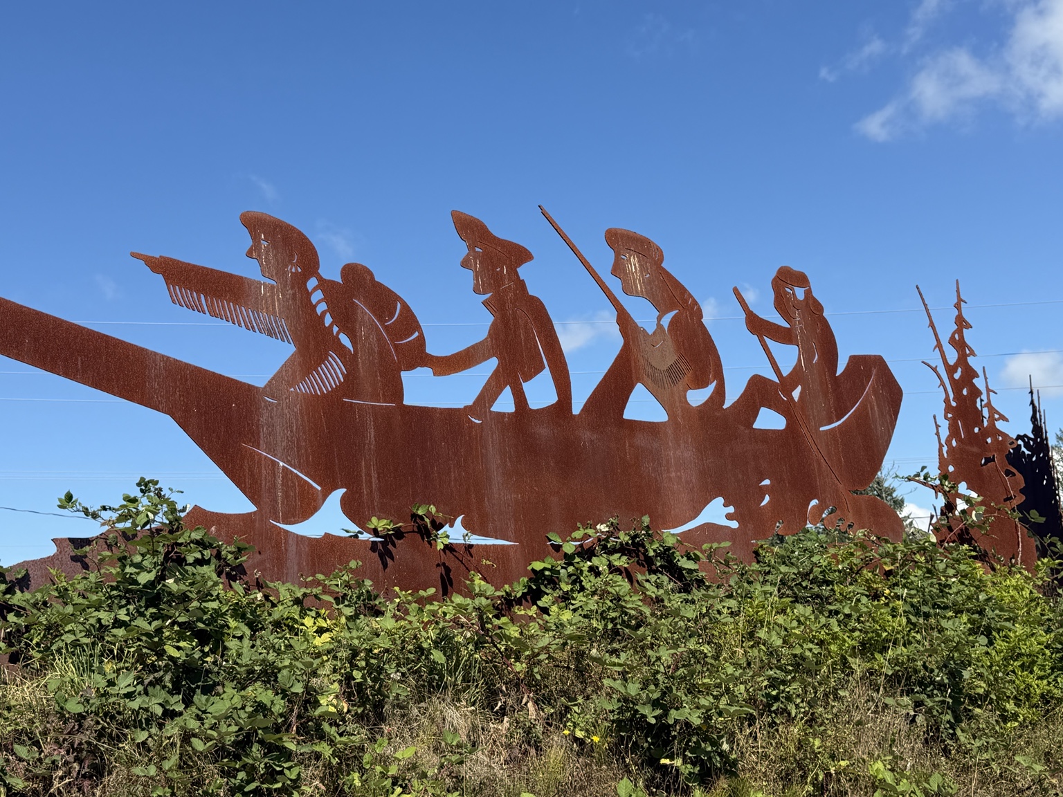 Rust-colored metal sculpture of people paddling a canoe beneath a clear blue sky